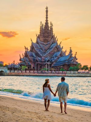 A diverse multiethnic couple of men and women visit The Sanctuary of Truth wooden temple in Pattaya Thailand at sunset on the beach