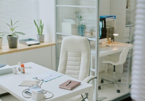 Bright and airy medical office with white furniture, green plants, and various medical equipment arranged on desk and in the room. Shelves and glass partitions adding to organized look